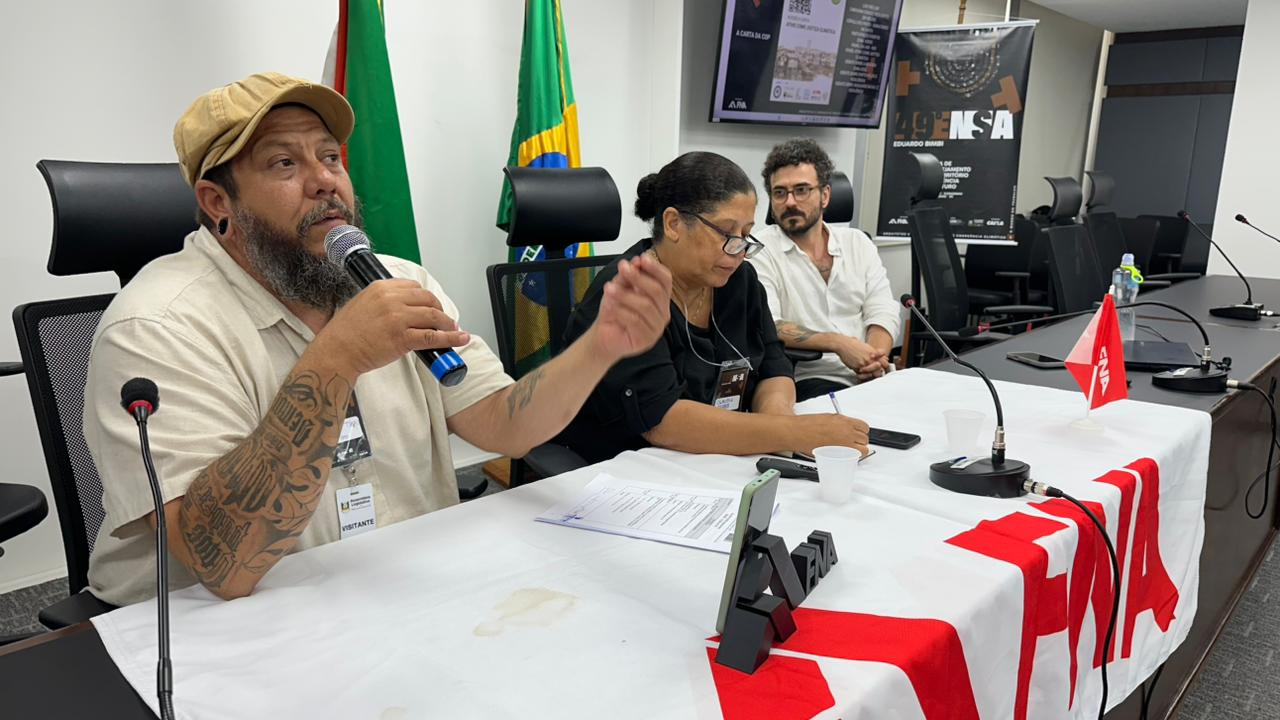 Filemon Tiago fala ao microfone numa mesa do ENSA, com as bandeiras do Brasil e do Rio Grande do Sul ao fundo